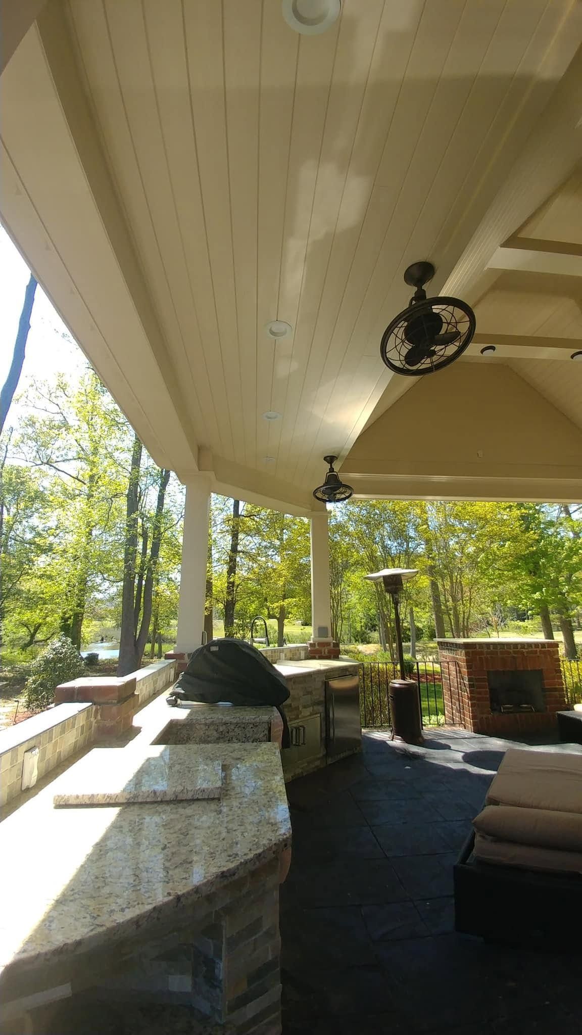 Covered outdoor patio with white ceiling, stone counter, cushioned seating, and trees beyond the railing