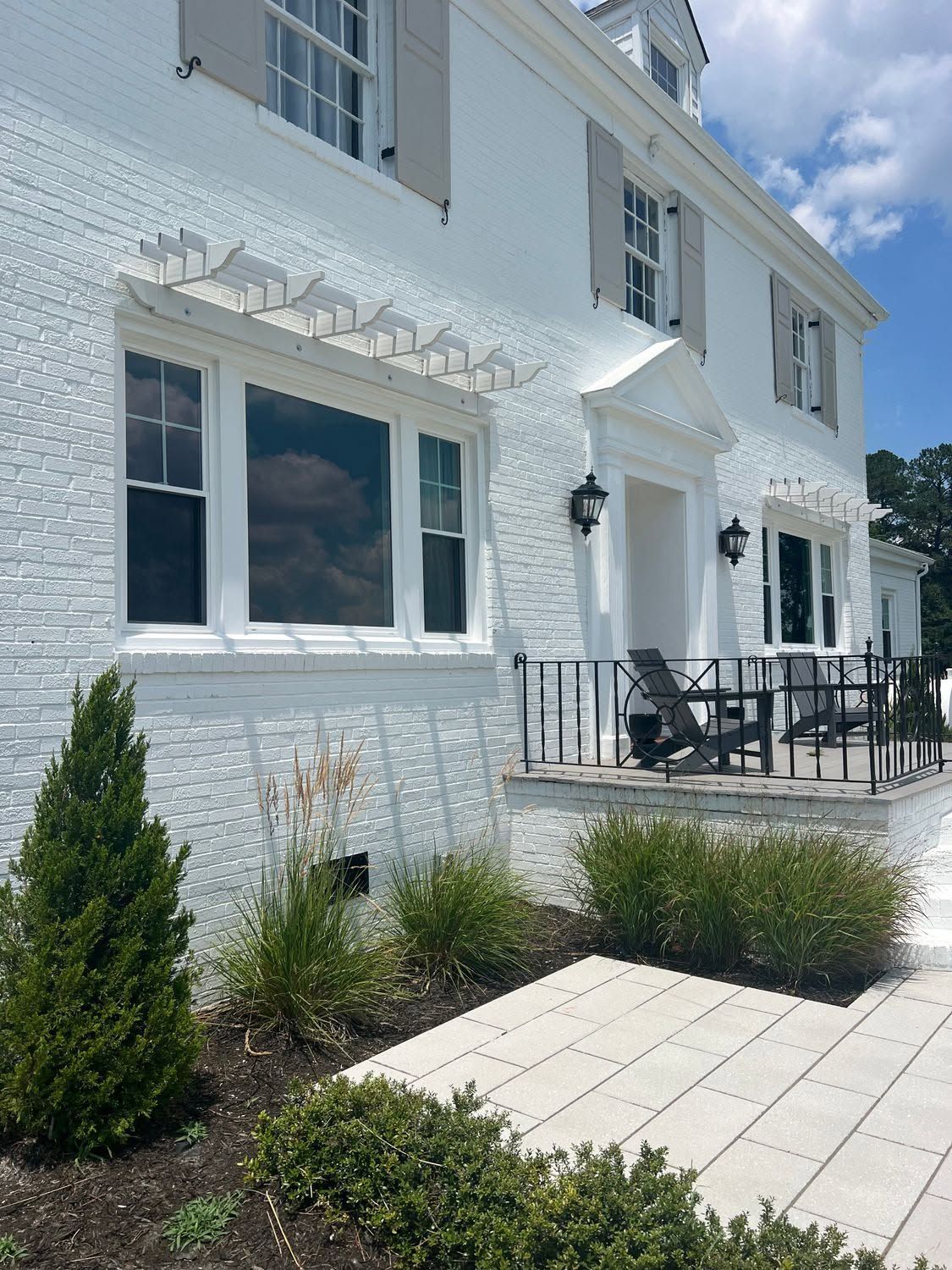 White house exterior with patio, black railing, and landscaped shrubs beside a paved walkway