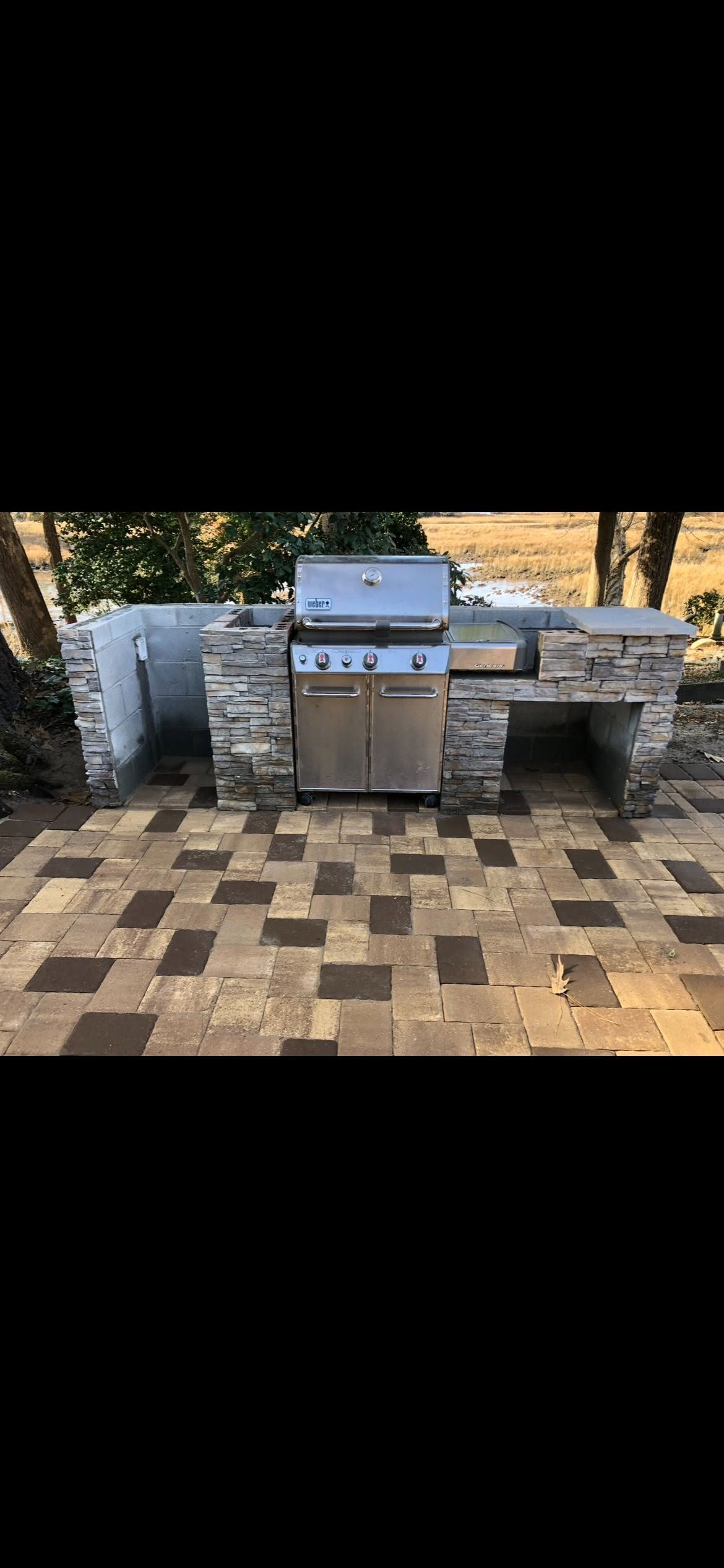Outdoor gas grill centered on a checkered stone patio, with a stone bench and wall behind it.