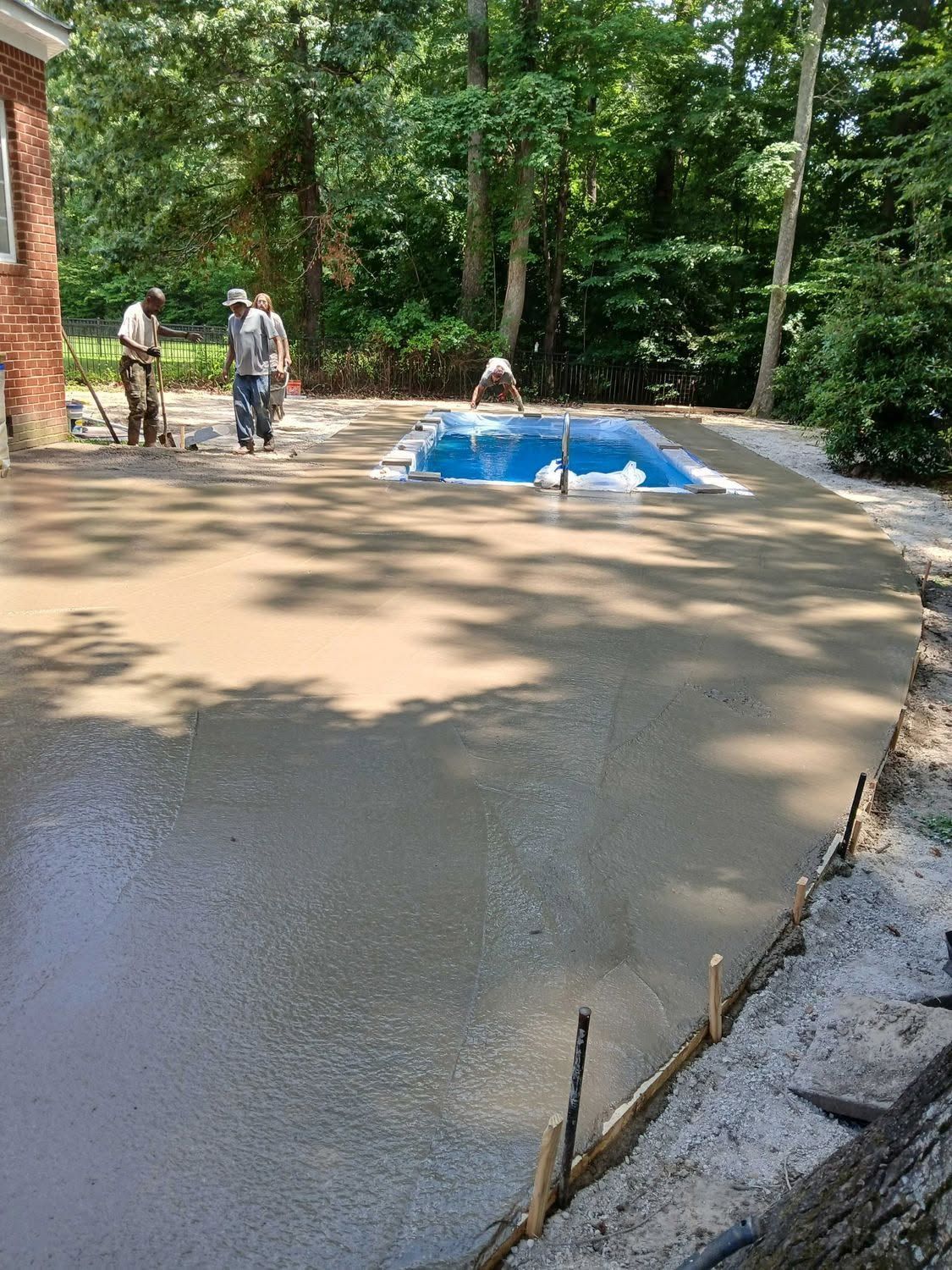 Workers smoothing fresh concrete driveway beside a house, with blue tarp and trees in the background