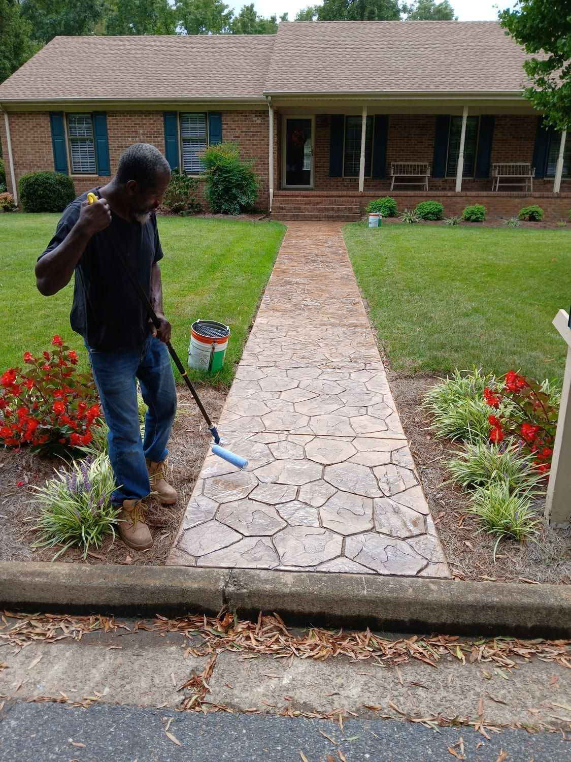 Person edging a front walkway in a suburban yard with red flowers and a brick house in the background