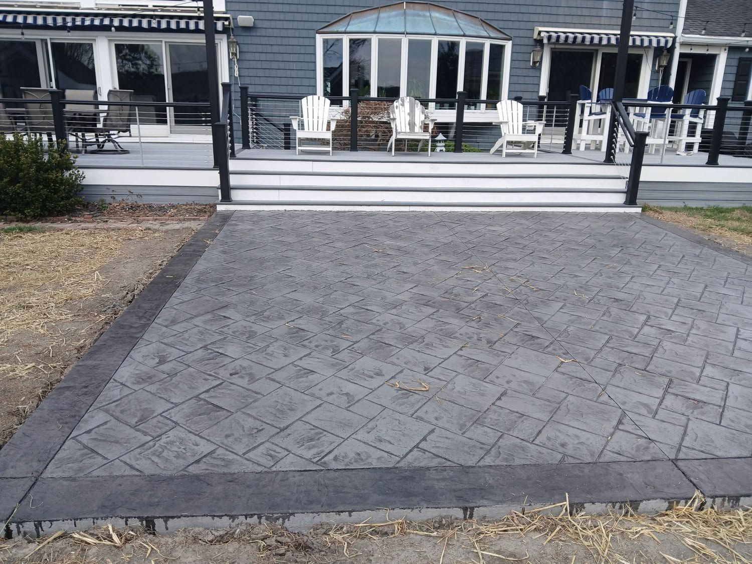 Paved stone patio leading to a house deck with white railing and outdoor chairs