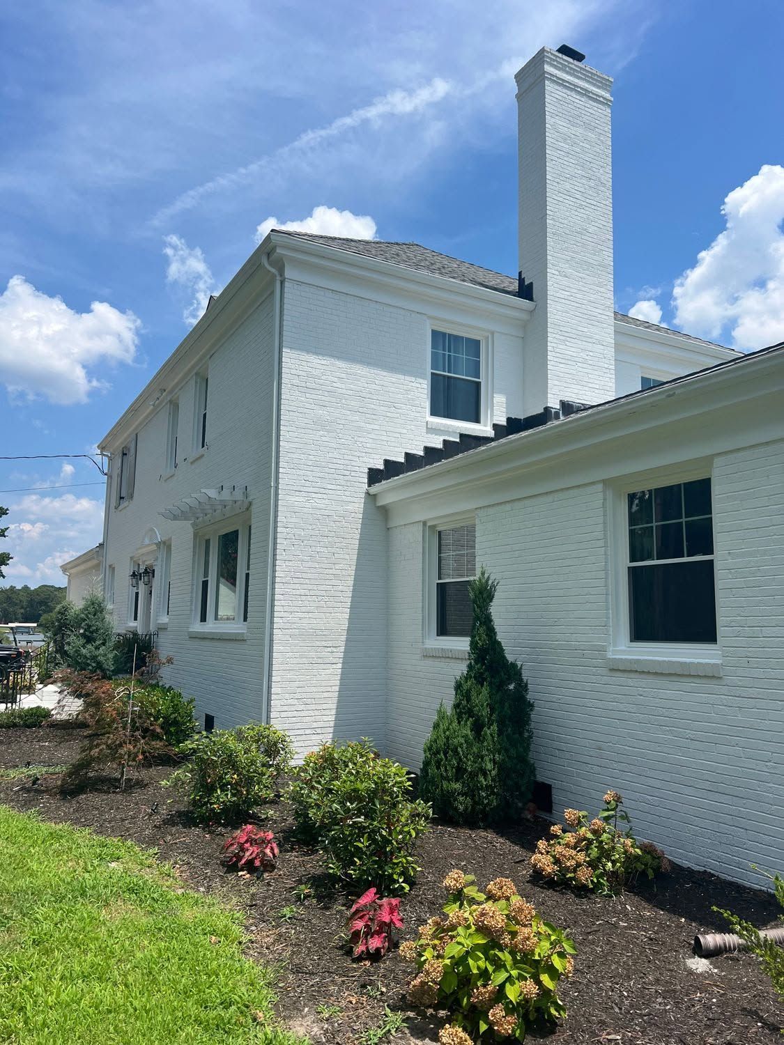 White two-story house with a tall chimney, black-trimmed windows, and a flower bed under a sunny blue sky