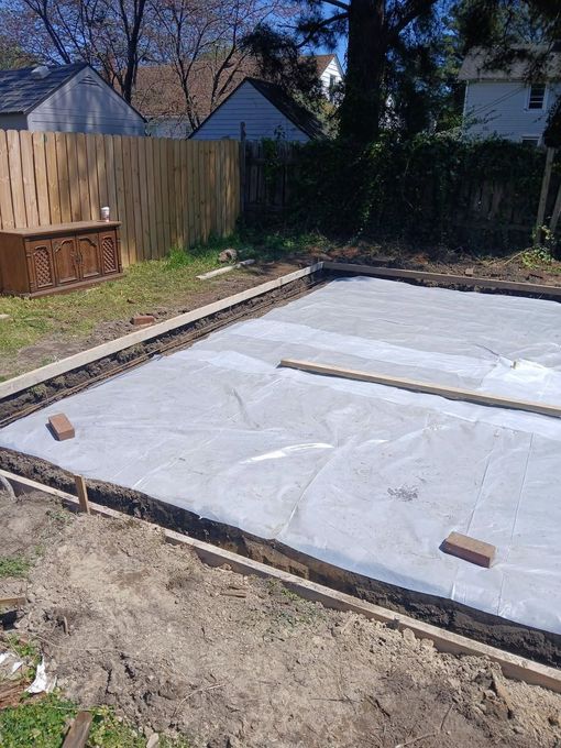 Backyard with a freshly covered rectangular ground tarp bordered by wood and bricks, near a fence and shed-like structures