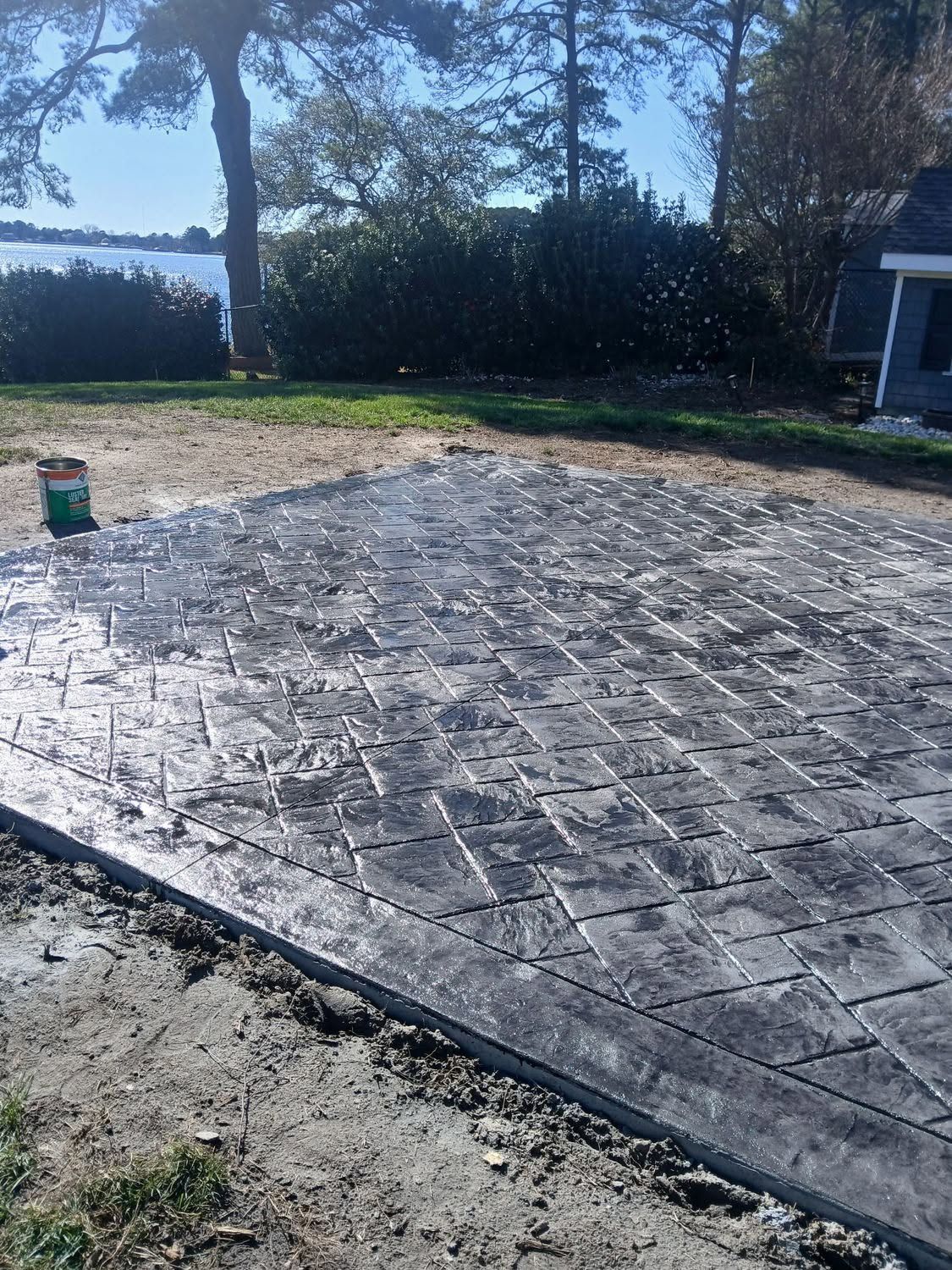 Stamped concrete patio under construction beside a yard and house, with a green bucket on the left.