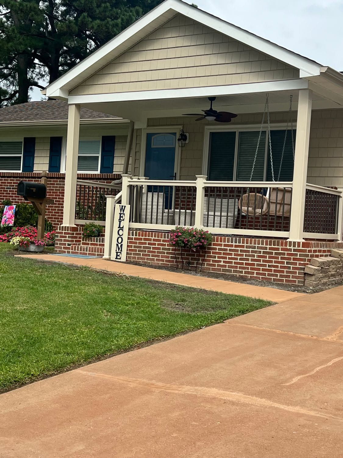 Front porch of a beige house with brick steps, white railing, and a curved driveway in front.
