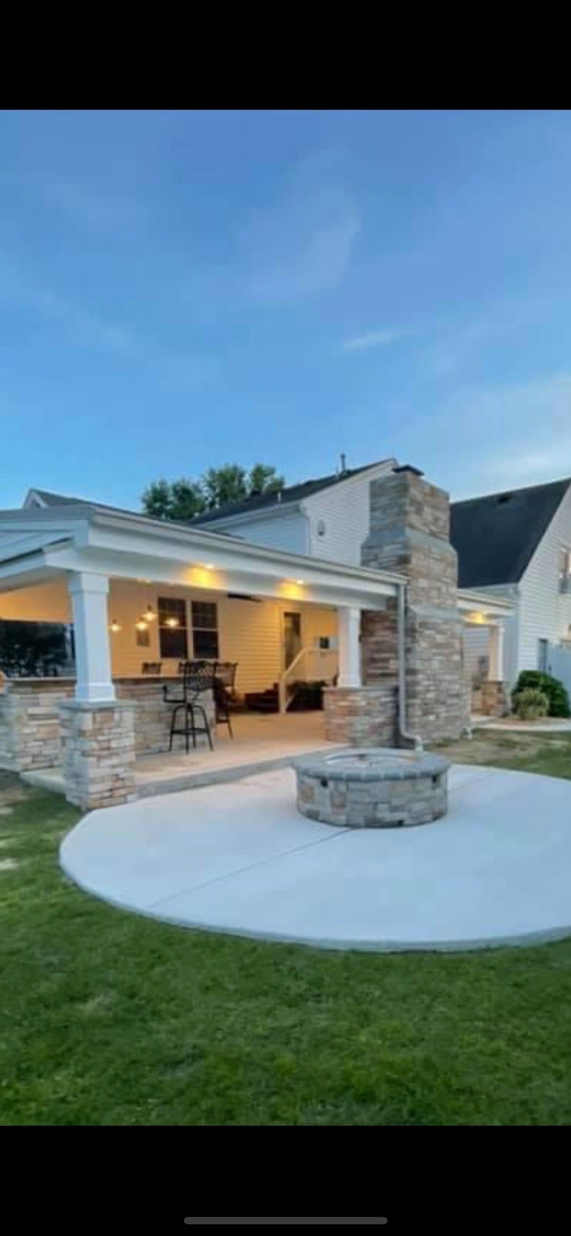Modern stone house patio with a lit covered porch, fire pit, and green lawn at dusk