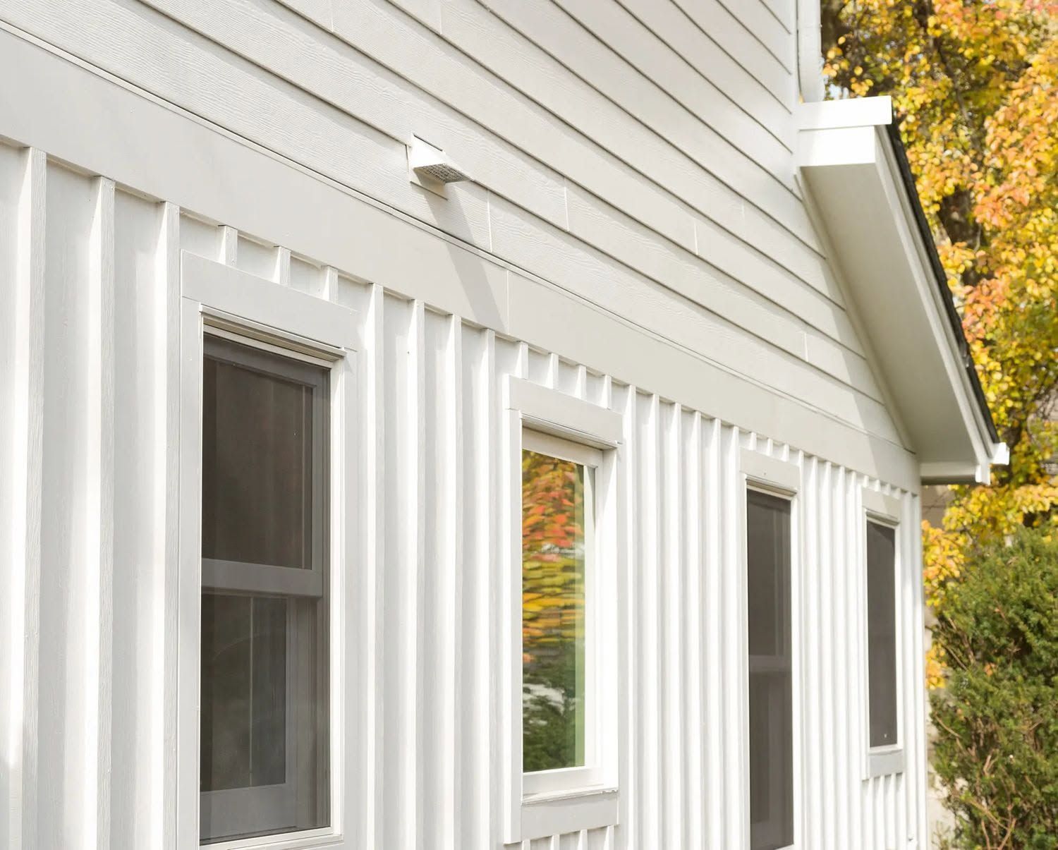 White house exterior with vertical siding, windows, and autumn trees in the background