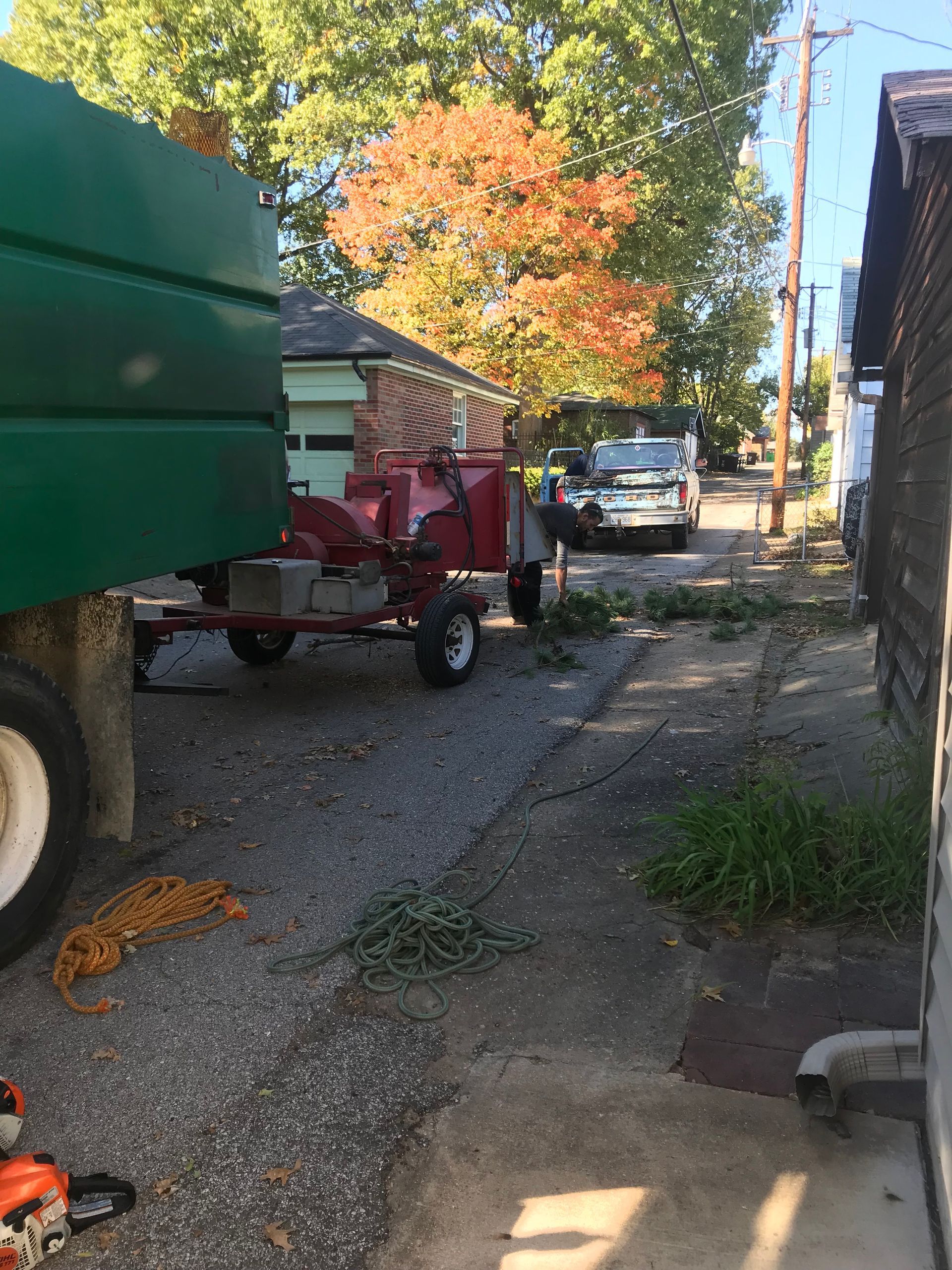 Gravel alley with a red trailer, green truck edge, and white van under autumn trees.