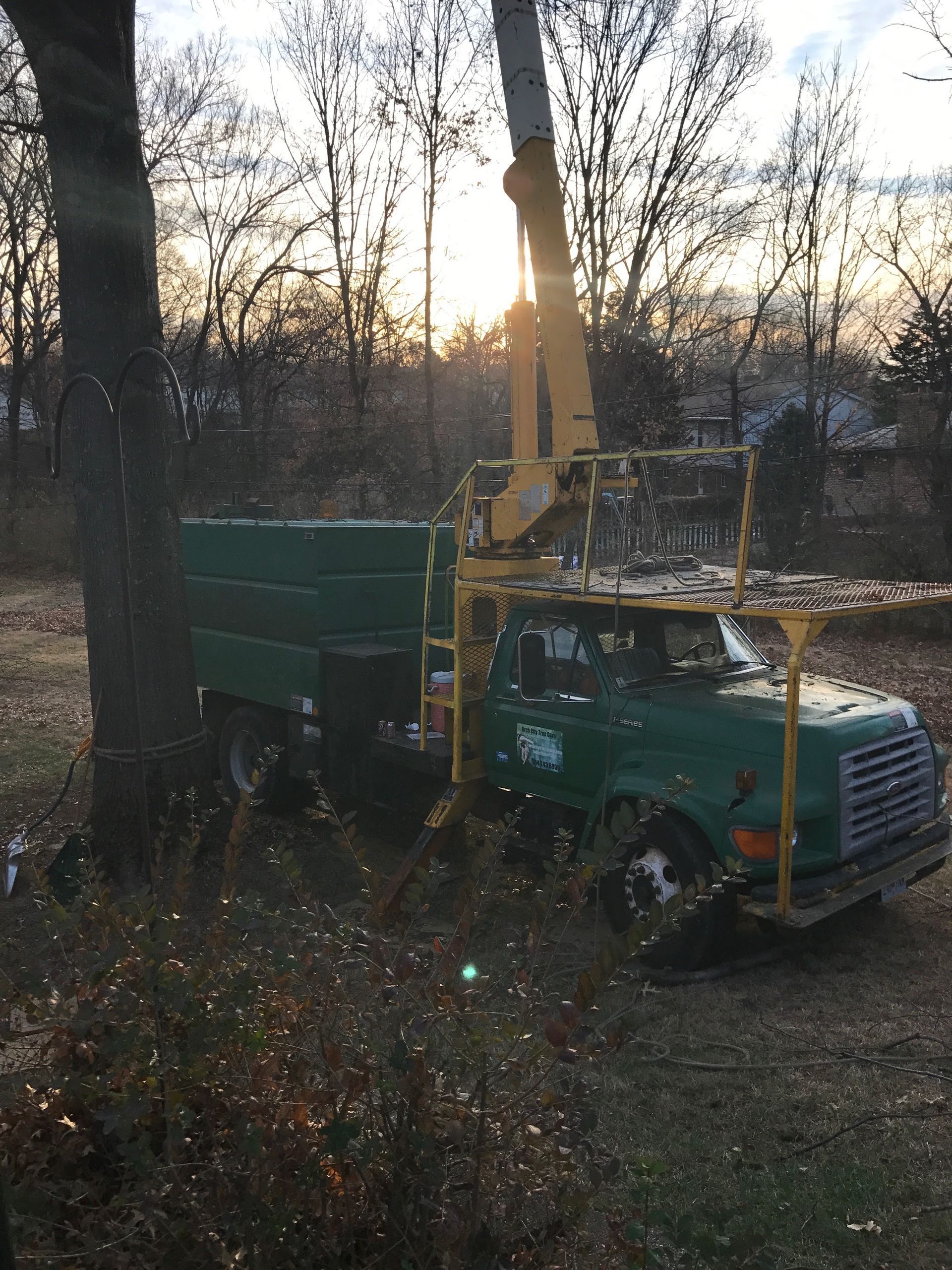 Green truck with a yellow crane parked in a wooded area at dusk