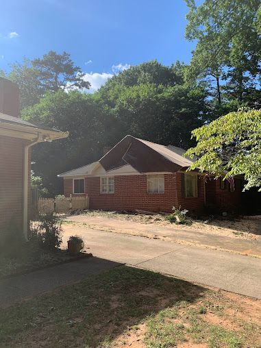 Red brick house with a sloped roof beside a driveway, surrounded by trees on a sunny day.