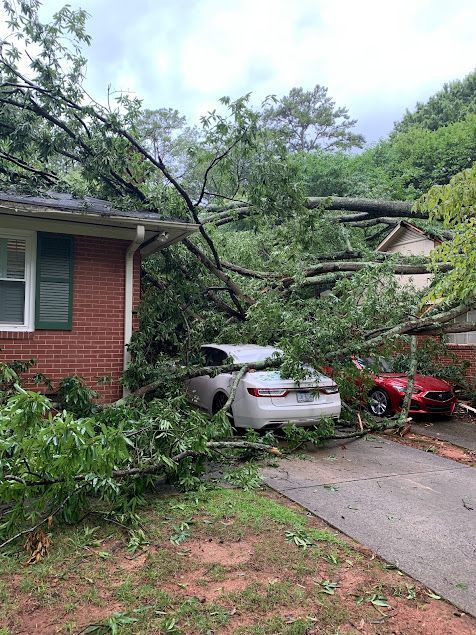 Storm damage in a driveway: fallen tree branches block a car beside a house.