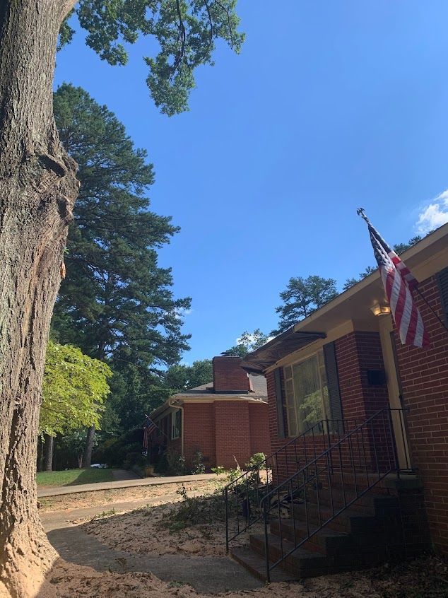 Brick house with steps and an American flag, shaded by a large tree under a blue sky