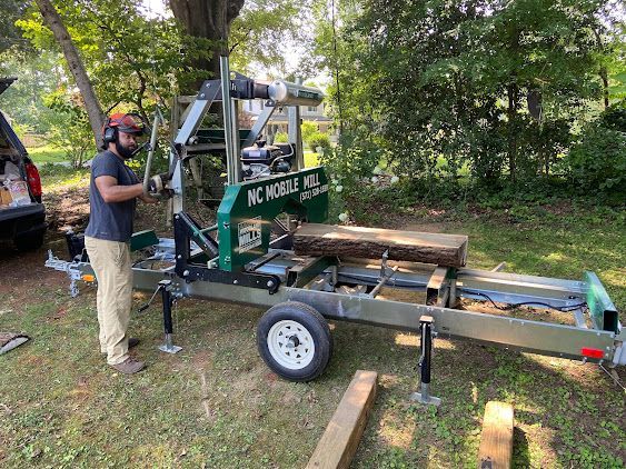 Man standing beside a green sawmill trailer in a wooded yard, with cut logs and lumber nearby