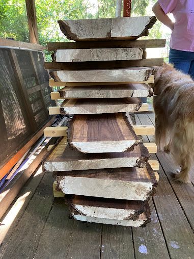 Stacked live-edge wood slabs on a deck, with a person and a dog partially visible beside them.