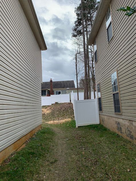 Narrow grassy passage between two houses, with a white fence gate and cloudy sky beyond.
