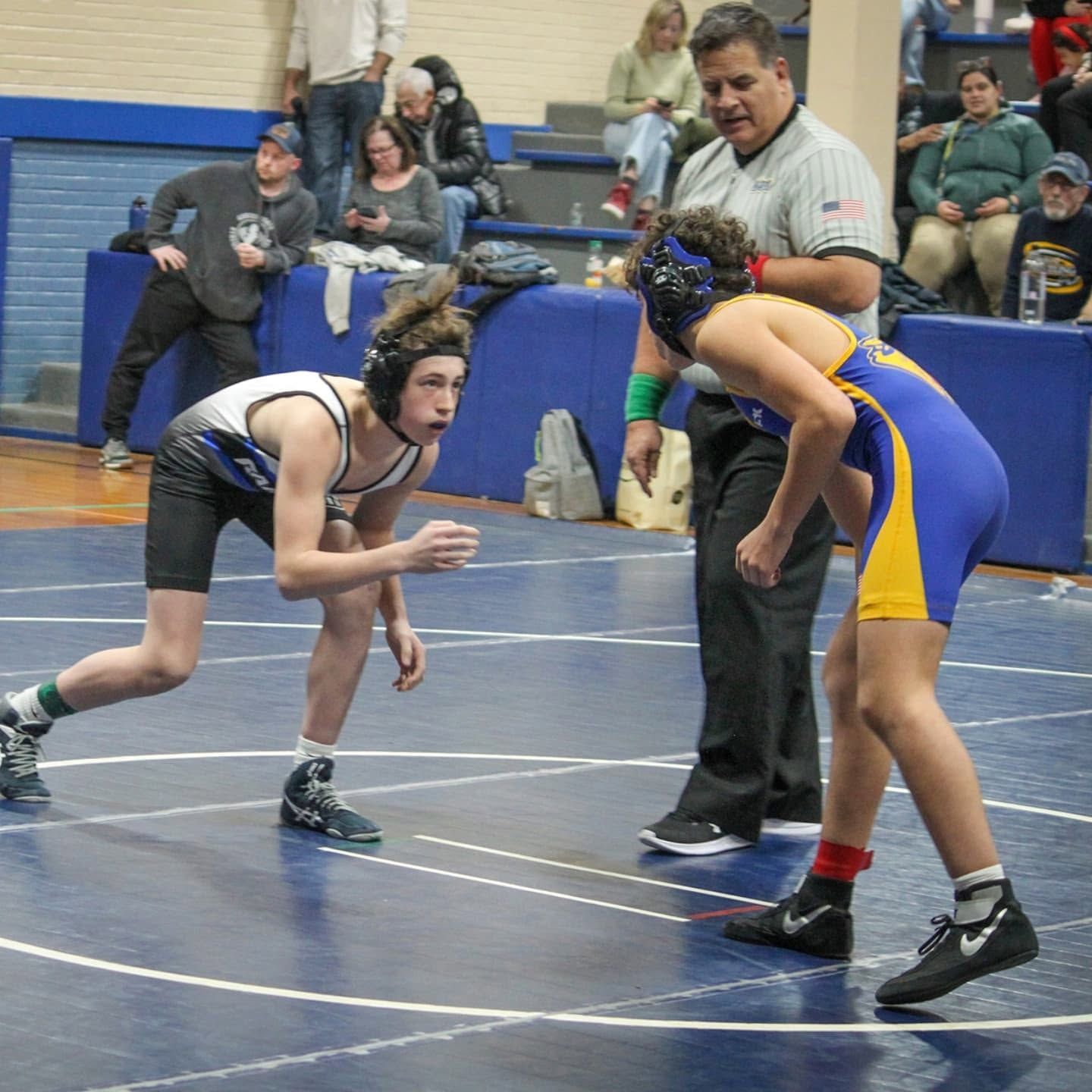 Two wrestlers in a match on a mat, crouched in stance with a referee nearby.
