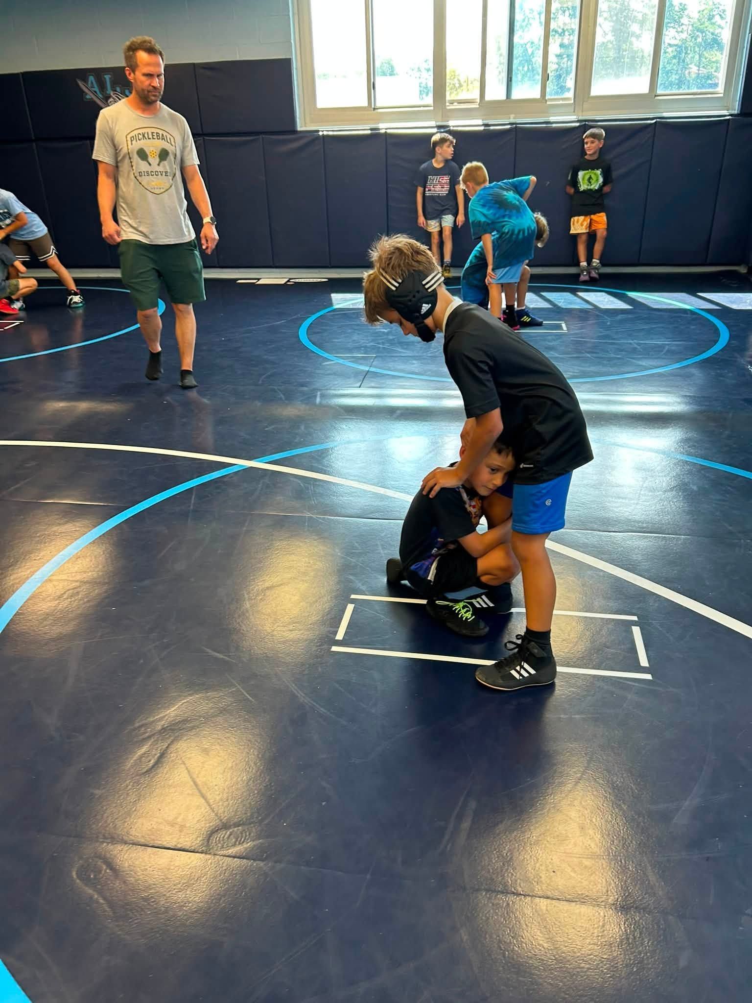 Youth wrestling practice in a gym, with a coach and children sparring on blue mats.