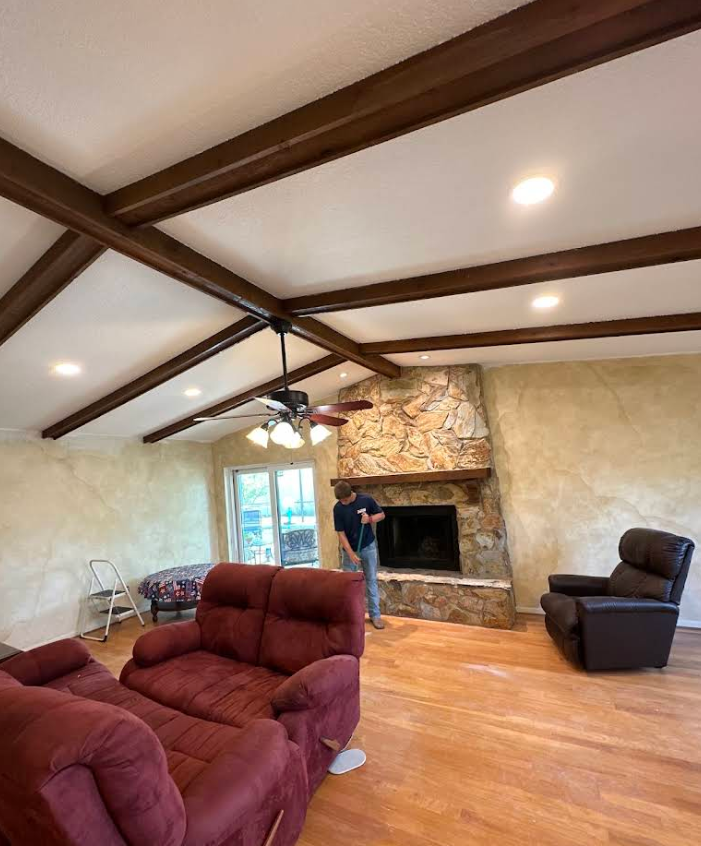 Spacious living room with wood beams, stone fireplace, red couch, recliner, and a person standing by the hearth