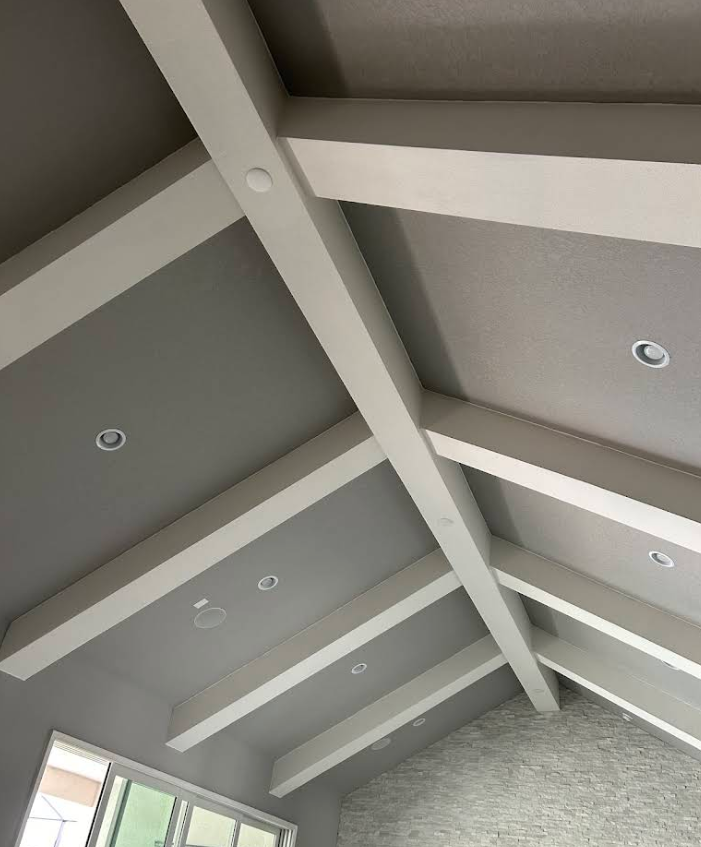 Vaulted ceiling with white beams and gray panels, with recessed lights above a stone wall.