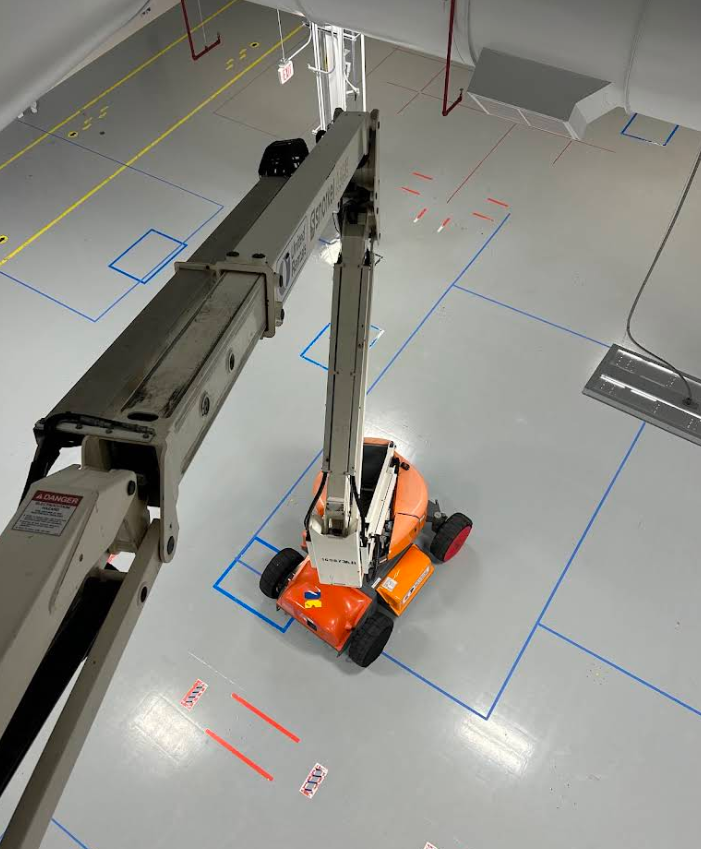 Orange robot arm in a lab, reaching toward a gray overhead beam on a marked floor.
