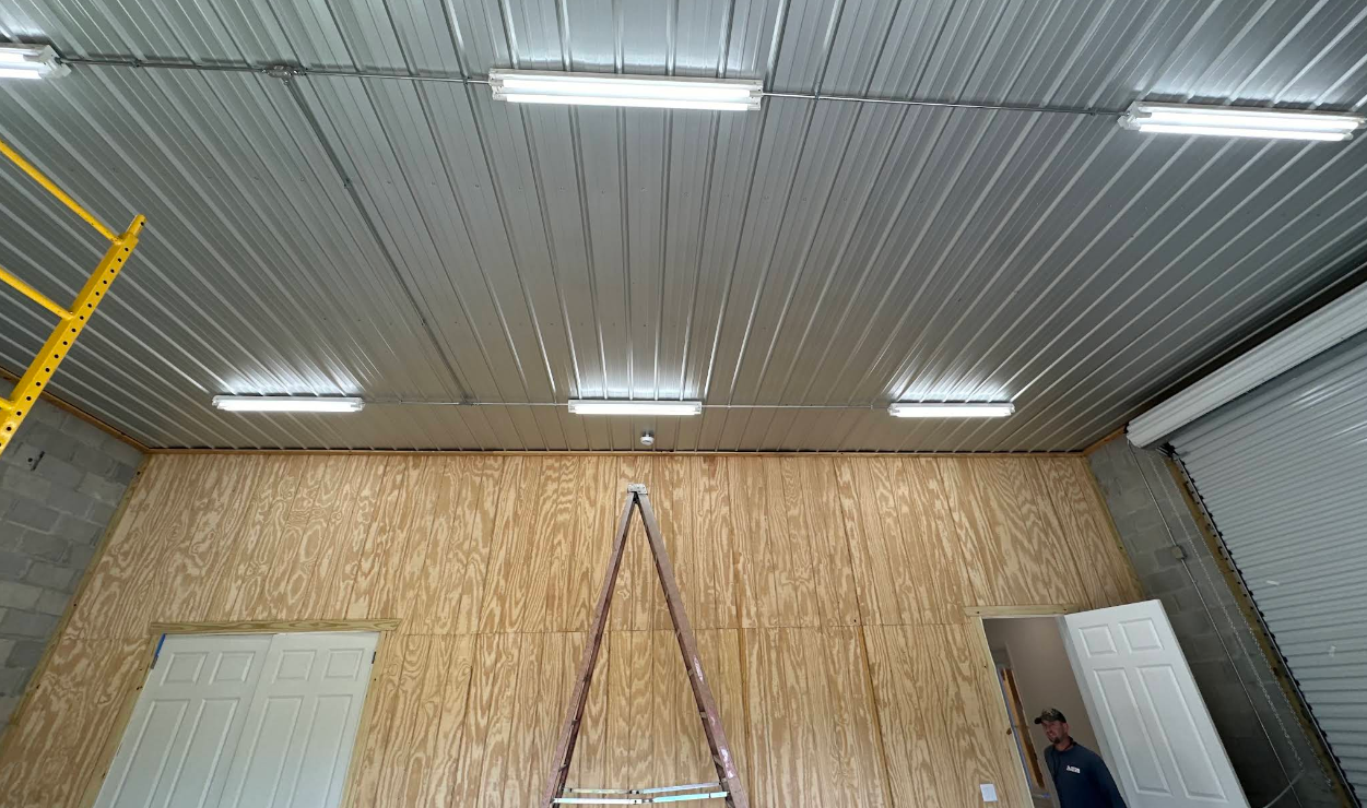 Warehouse interior with corrugated metal ceiling, wooden wall, lights, and ladder against the wall.