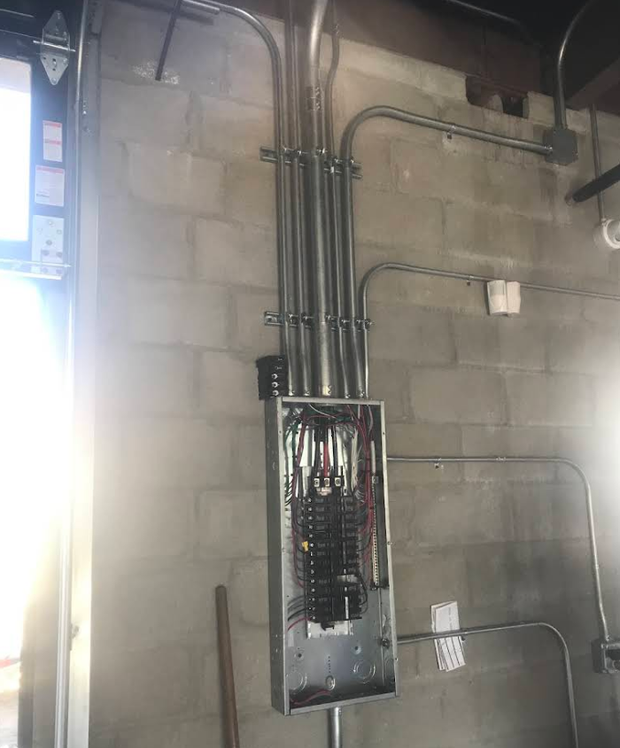 Open electrical breaker panel on a cinderblock wall with conduit pipes above it