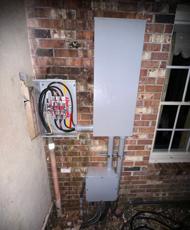 Electrical panels mounted on a brick exterior wall beside a window, with exposed wiring and conduits.