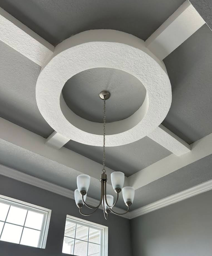 Gray coffered ceiling with a round recessed center, hanging chandelier, and crown molding above windows.
