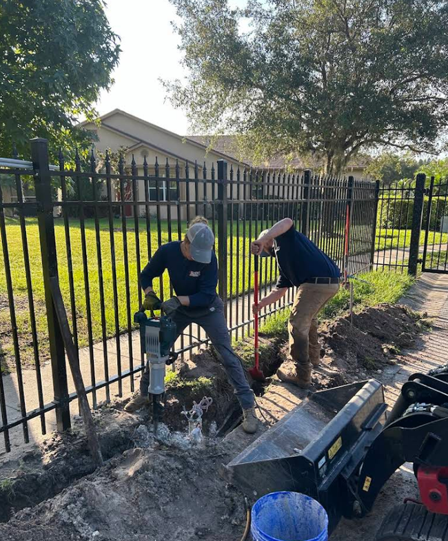 Two workers digging near a black fence beside a sidewalk, with tools and trenching equipment.