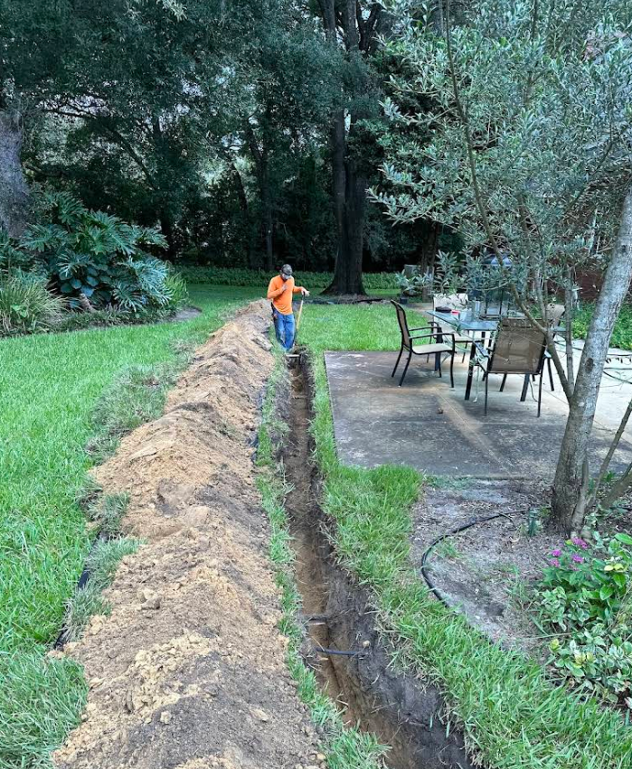 Person in orange standing by a long trench beside a grassy yard with patio furniture and trees