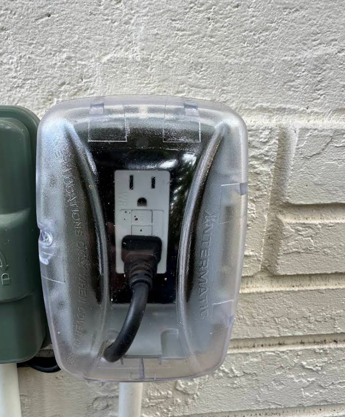 Covered outdoor electrical outlet with a plugged-in black cord on a brick wall