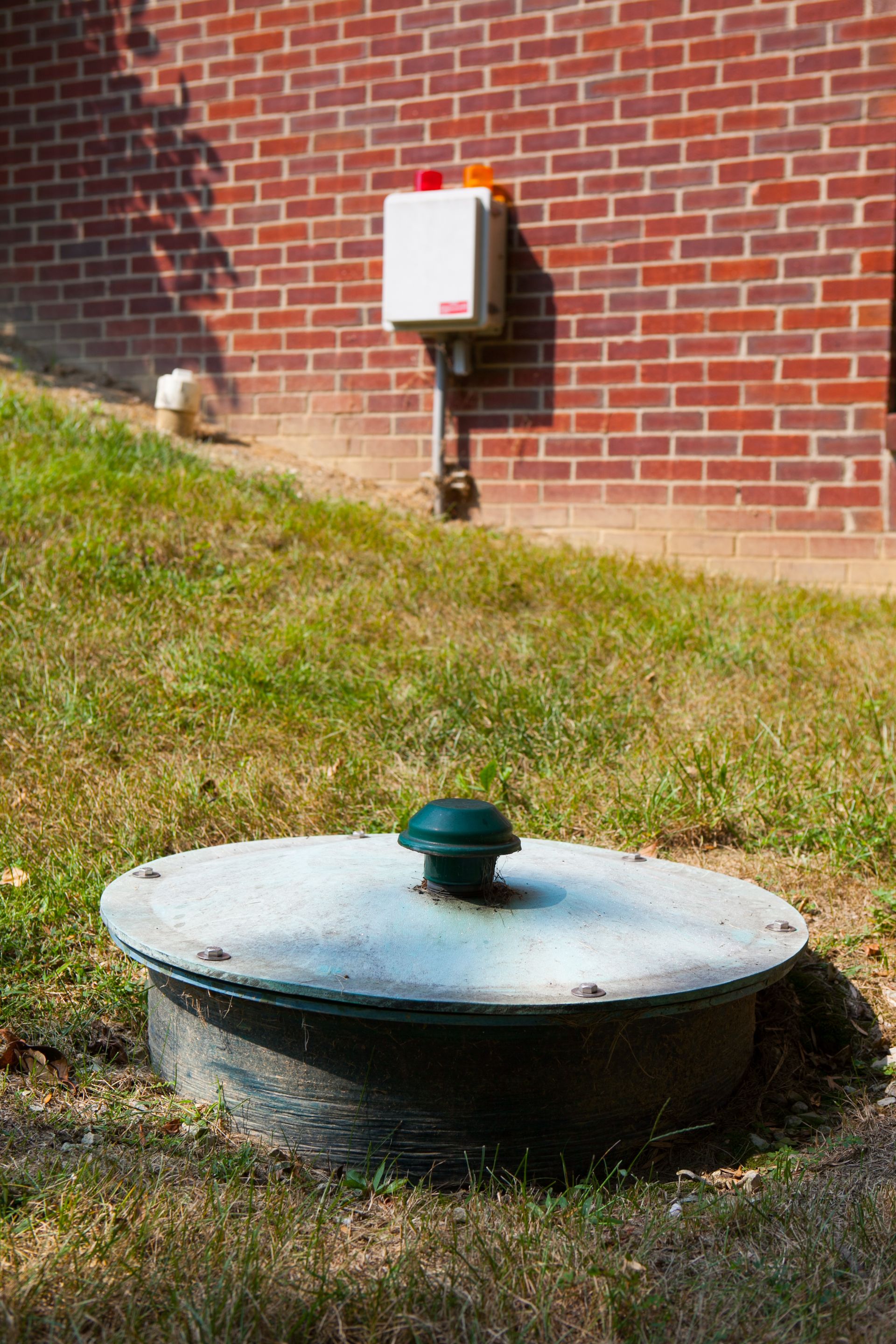 Green utility lid beside a brick building and grassy slope