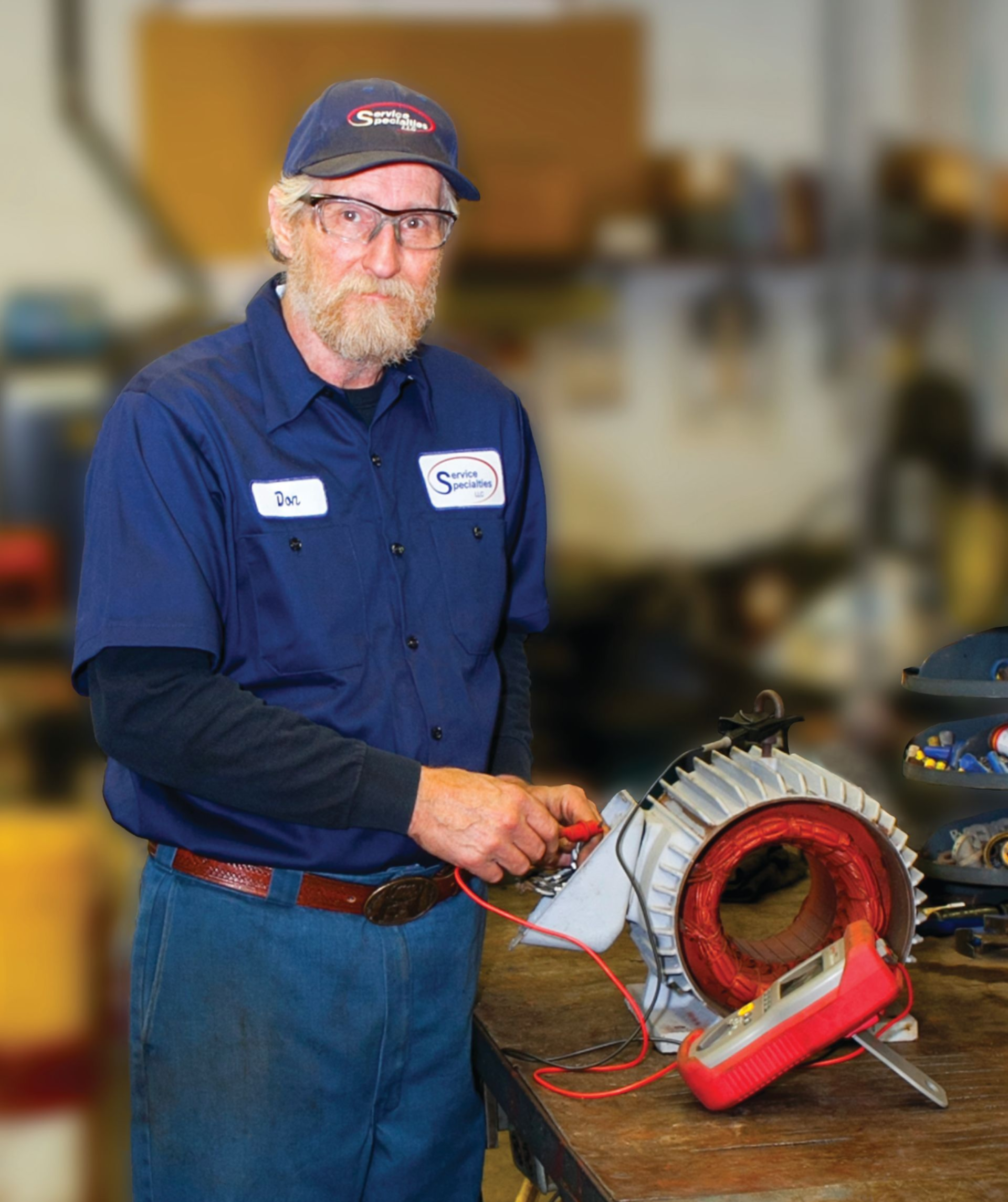 Worker in blue uniform holding a red power tool beside a large metal coil in a workshop
