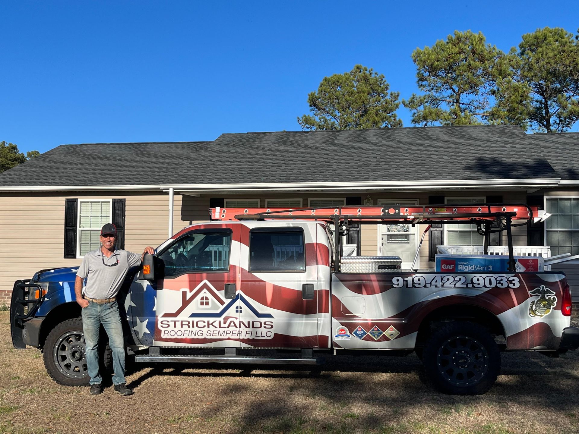 A person standing next to a truck wrapped in an American flag design with the Strickland's roofing logo in front of a house.