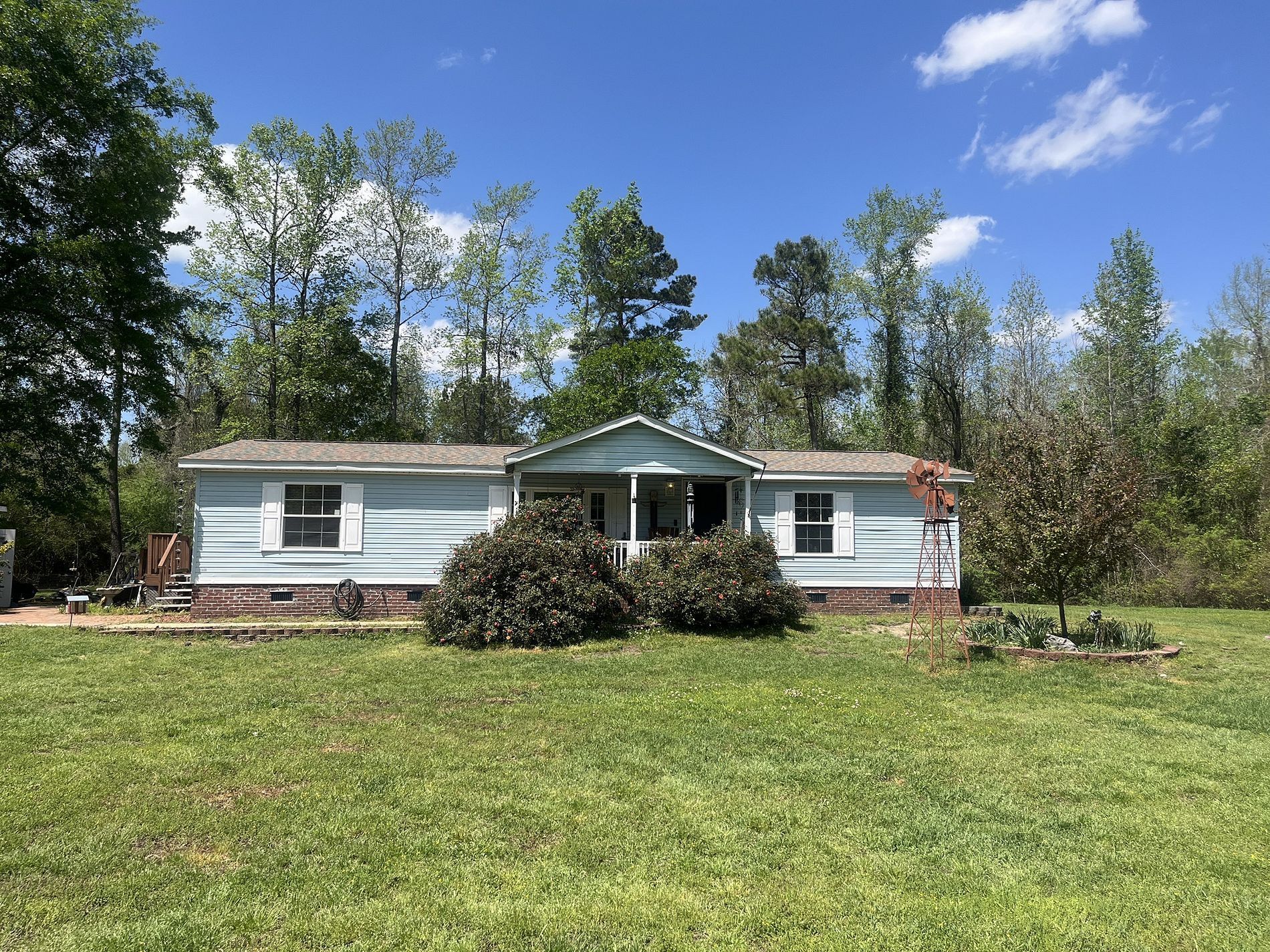 A light blue manufactured home with a covered porch sits on a grassy lawn surrounded by trees under a blue sky.