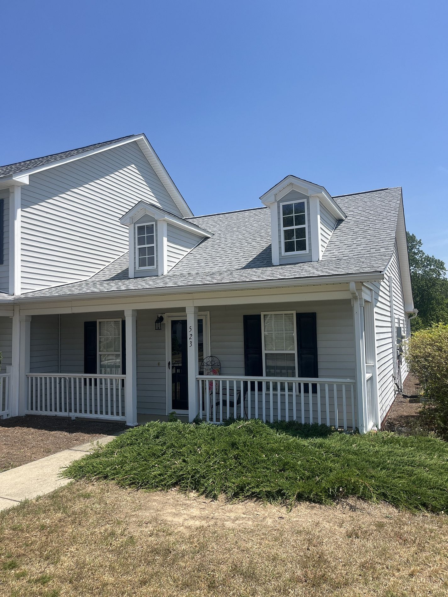 A light gray two-story house with a front porch, white railings, dark shutters, and two dormer windows.
