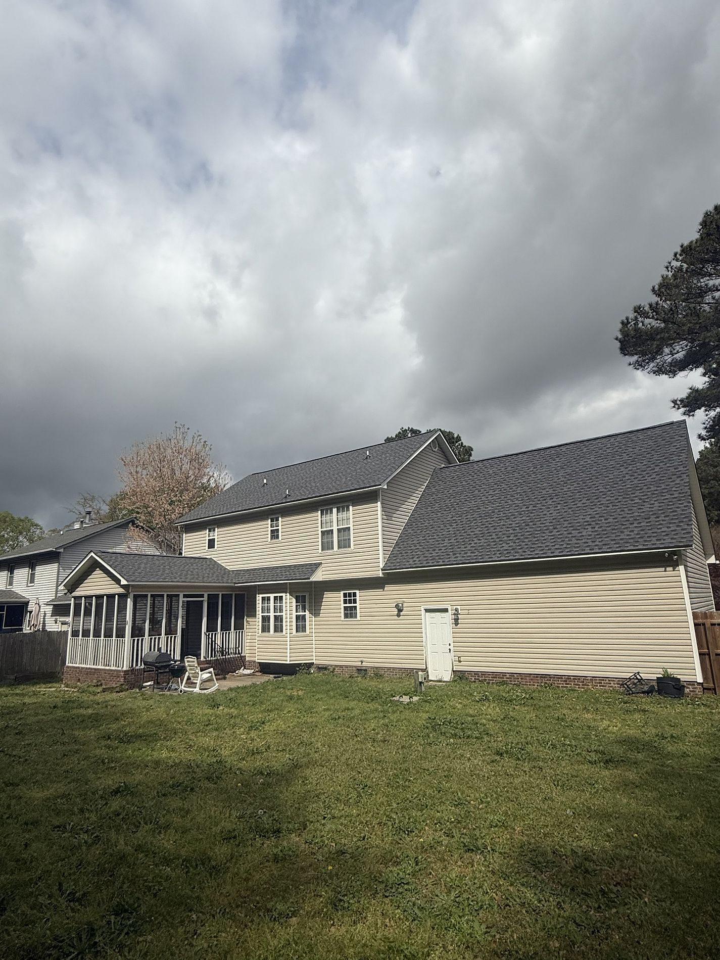 A tan, two-story house with a newly installed dark gray shingle roof under a cloudy sky.