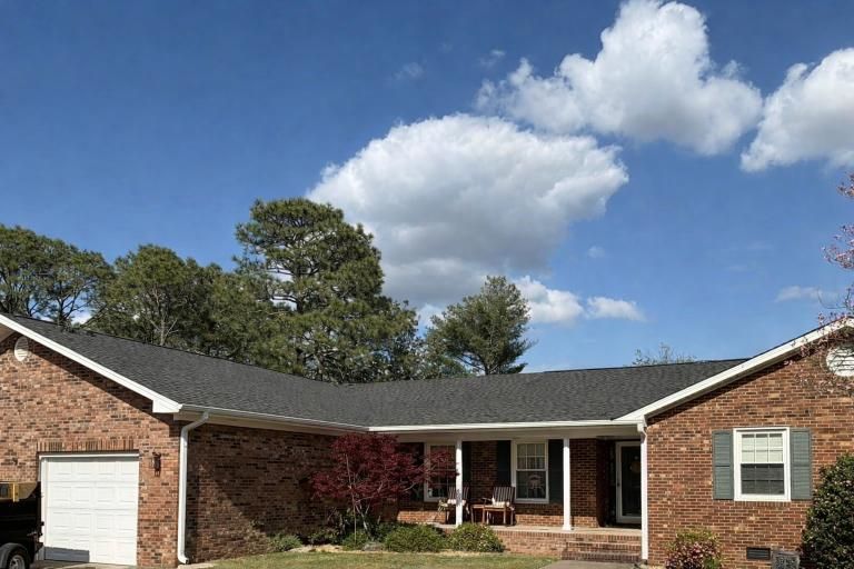 A single-story brick house with a dark shingled roof, attached garage, and front porch under a bright blue sky.