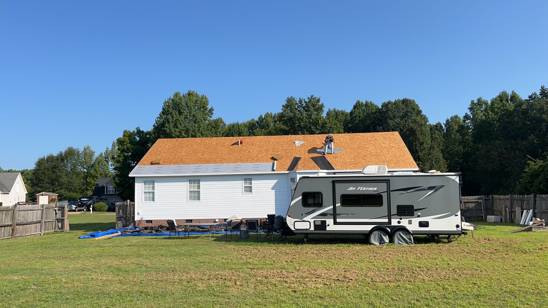 A white house with a newly exposed wooden roof sits on a grassy lot next to a gray travel trailer under a clear blue sky.