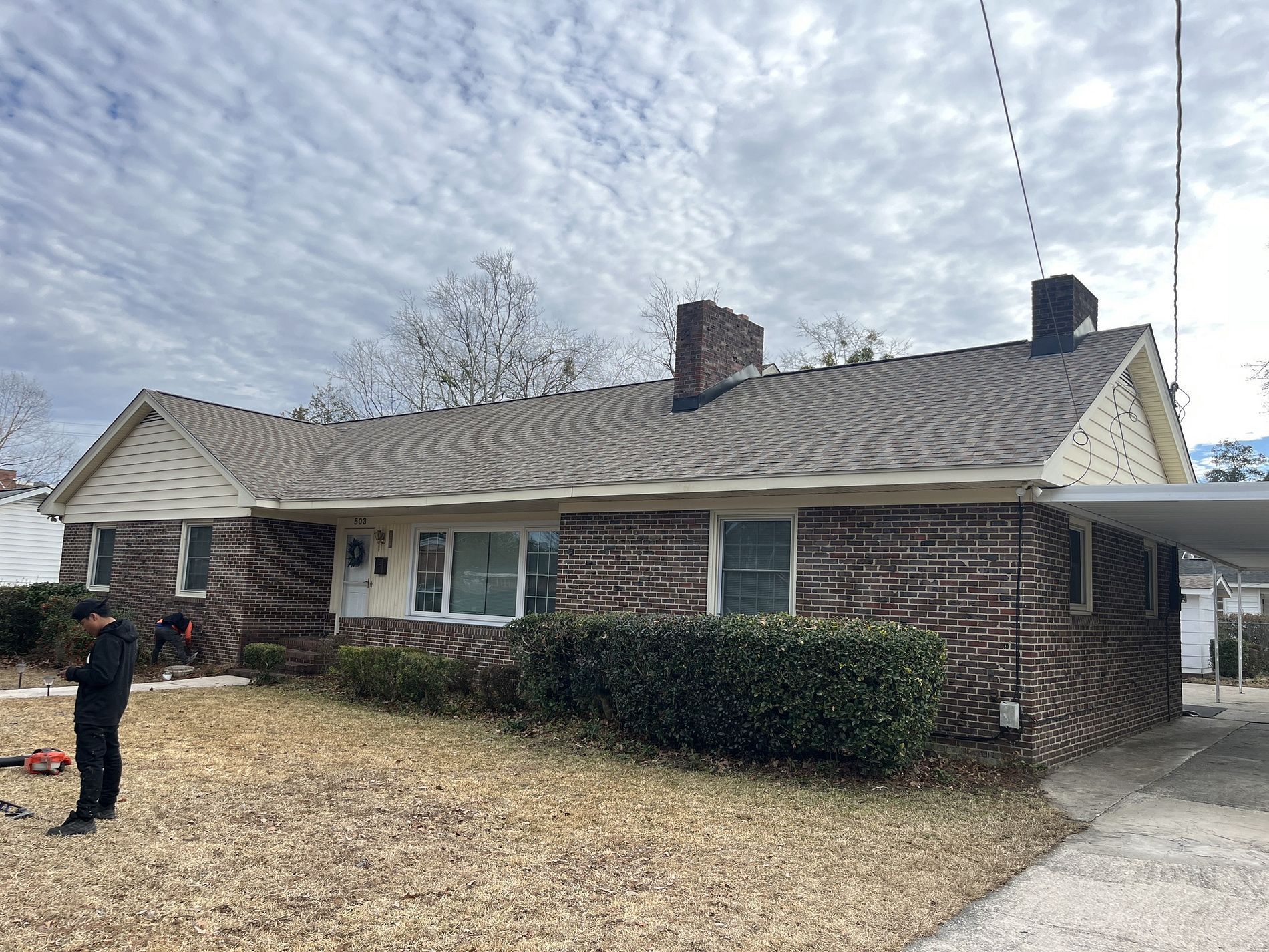 A man is standing in front of a brick house with a carport.