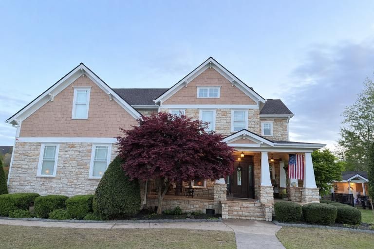 A two-story suburban home with tan stone and beige siding, a large burgundy tree in front, and an American flag.