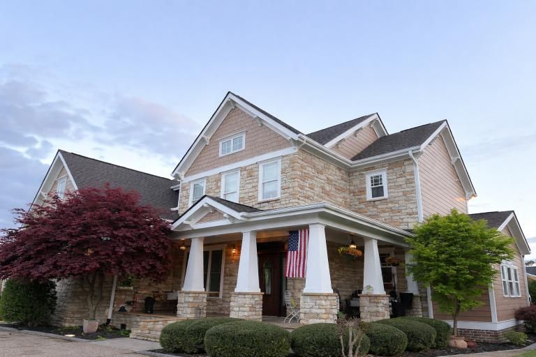 A two-story tan stone and siding house with a covered front porch, white pillars, a hanging American flag, and trees.