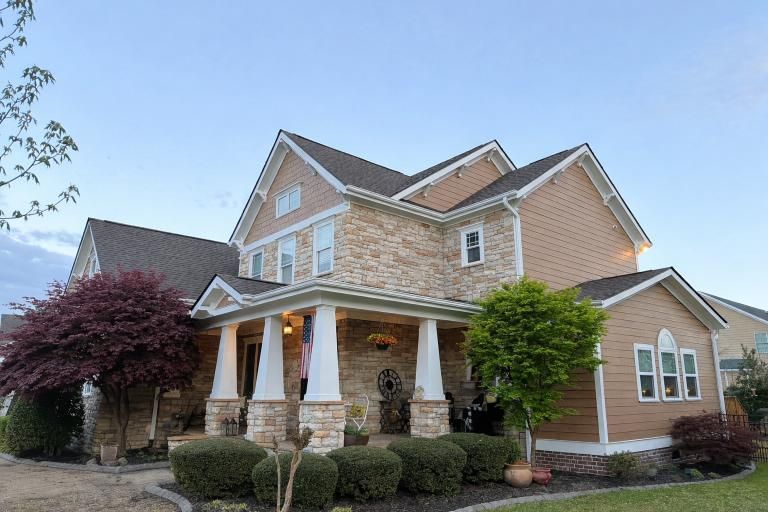 A two-story suburban home with tan siding, stone facade, covered front porch with white pillars, and landscaped shrubs.