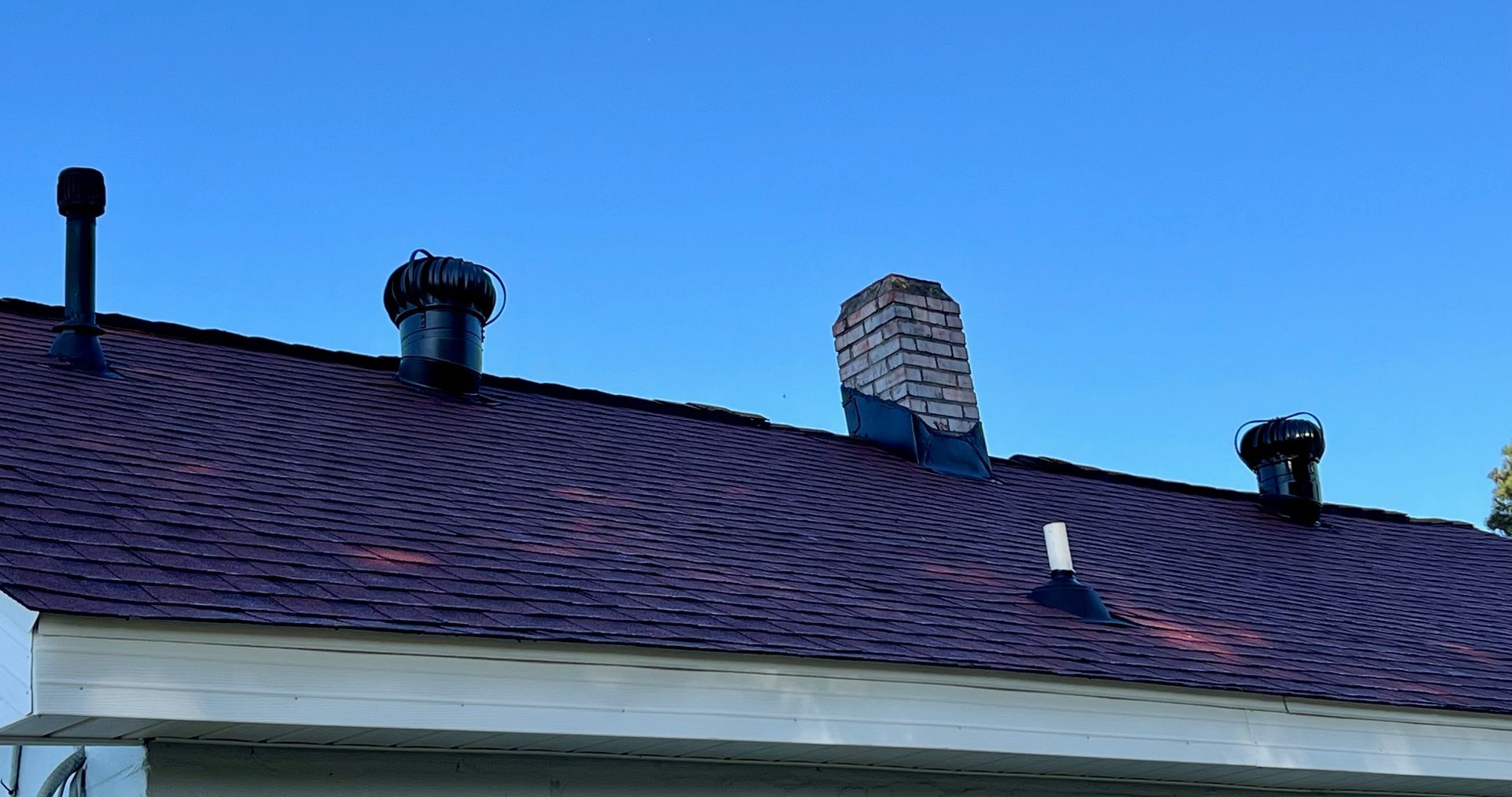 A residential roof with dark shingles, two black vents, a white brick chimney, and a small white pipe against a blue sky.