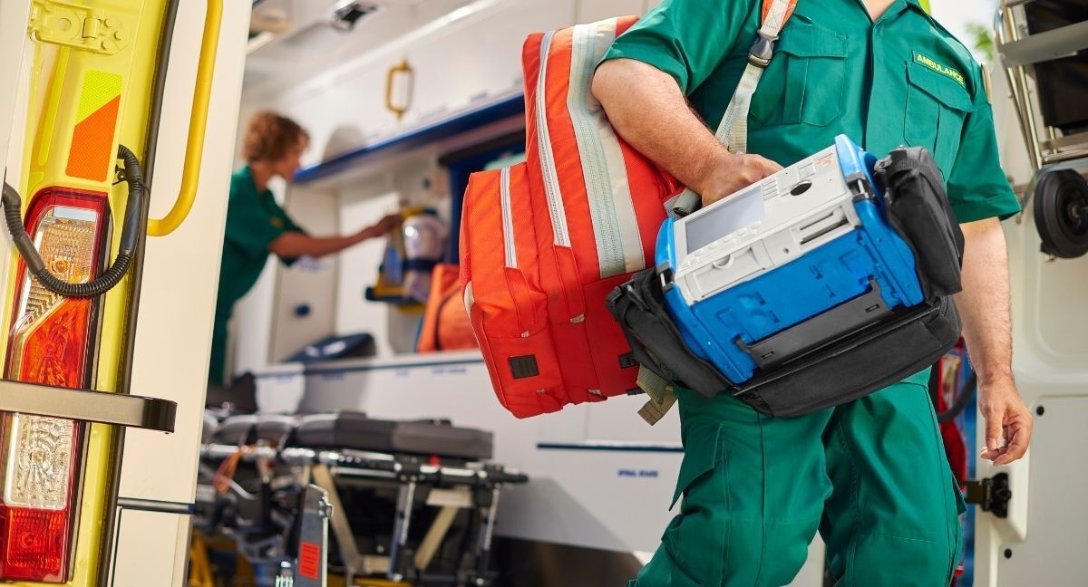 A paramedic is carrying a bag and a device in front of an ambulance.