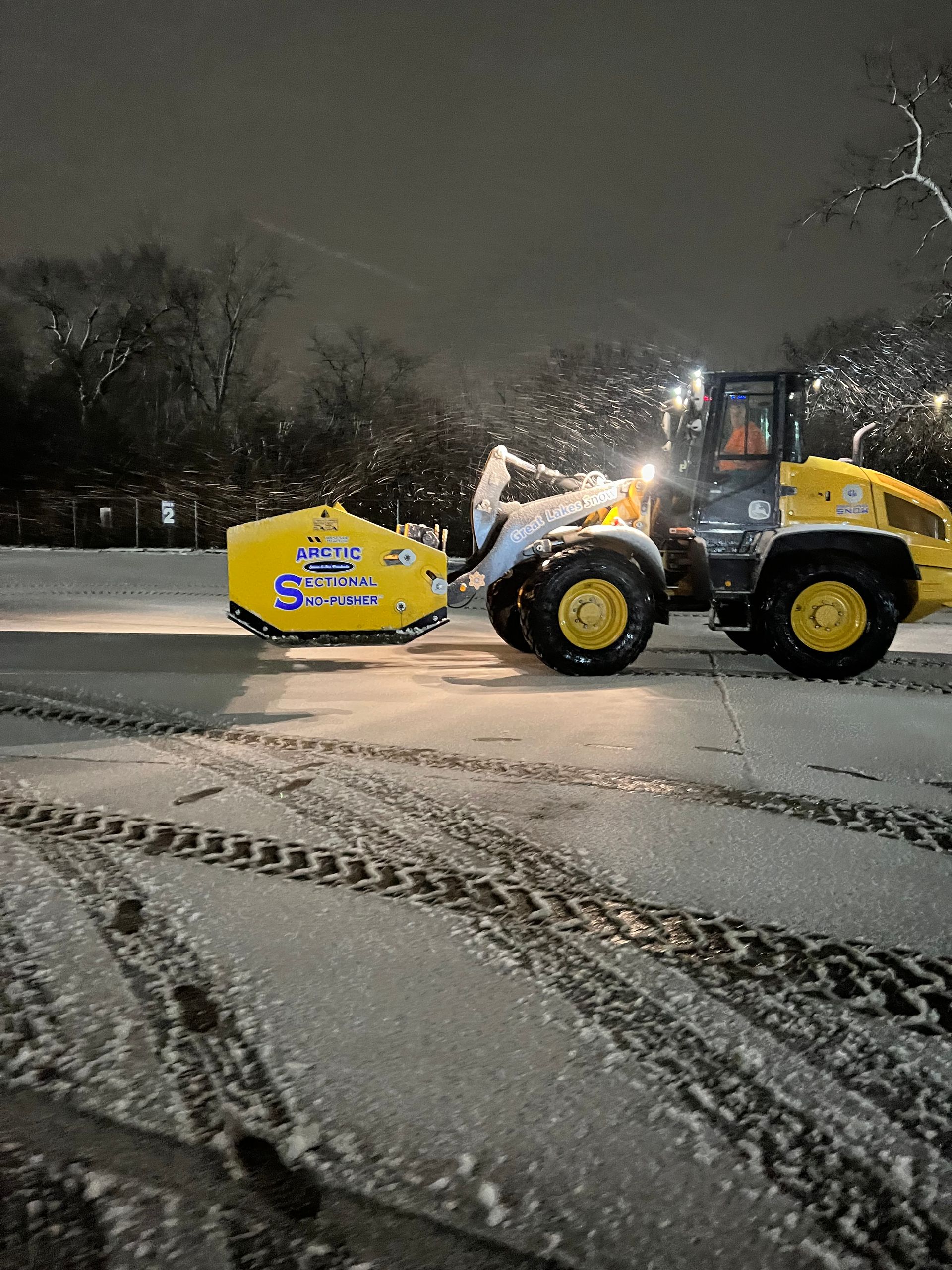 A Great Lakes Snow John Deere loader with pusher box clearing a commercial parking lot.