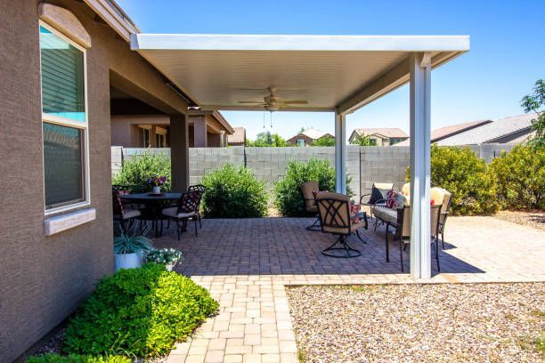 Covered patio with potted plants, outdoor chairs, and a brick paver seating area beside a house.