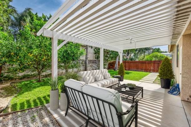 Covered patio with white pergola, cushioned seating, potted plants, and a fenced green backyard.