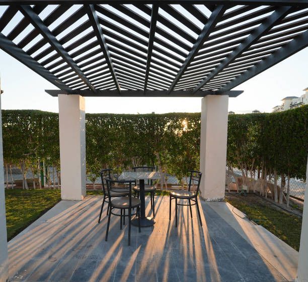 Outdoor patio with metal pergola, round table, and chairs on a sunlit deck beside trimmed hedges